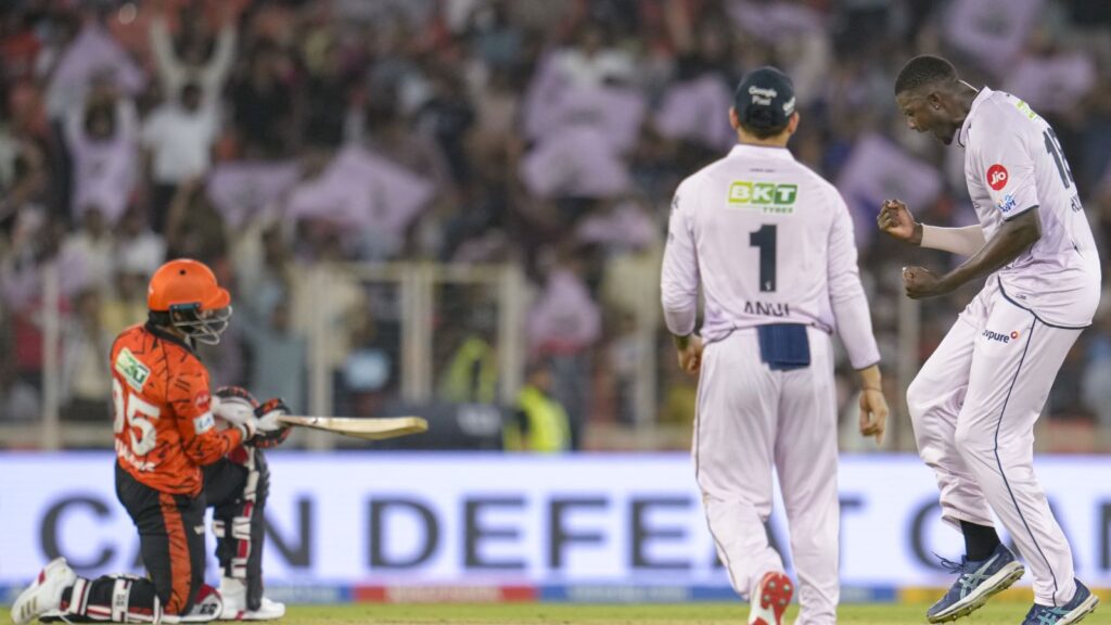 A cricket player in orange pads and helmet prepares to bat during a match at a stadium.