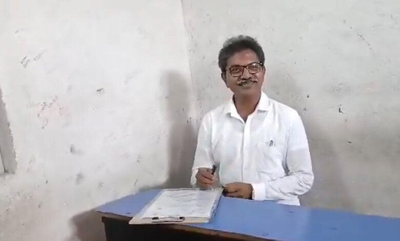 A person smiling and sitting at a desk with papers, wearing glasses and a white shirt.