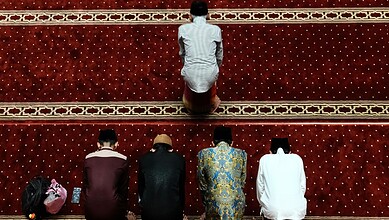 A Muslim man praying on a red prayer mat with four other men in a mosque.