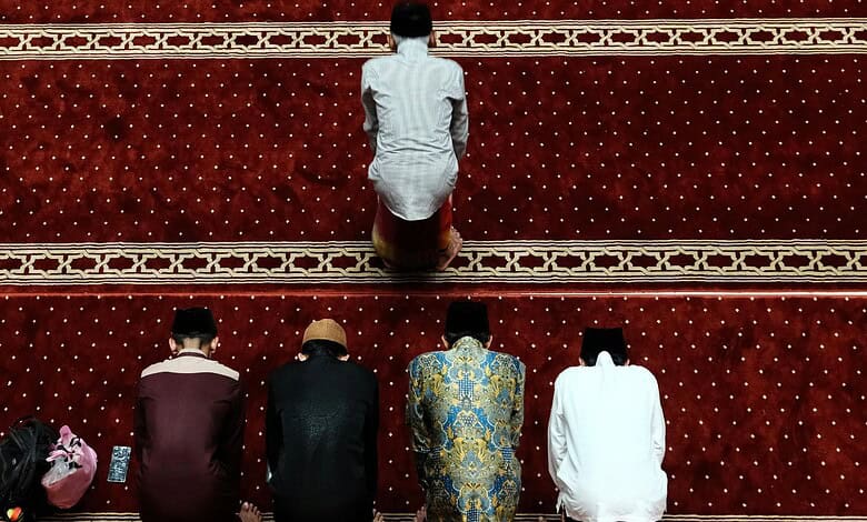 A Muslim man praying on a red prayer mat with four other men in a mosque.
