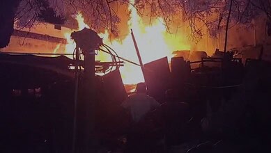 A firefighter battling a large blaze with intense flames and smoke in the background.