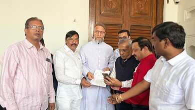 Group of Indian men in formal attire discussing documents indoors, in front of wooden door.