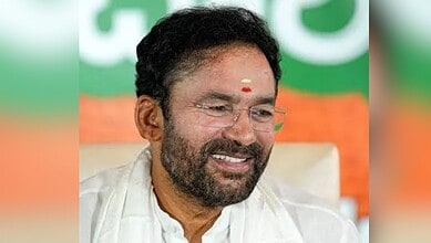 A man with glasses and a beard smiling, wearing a white traditional outfit, in front of a green and orang.