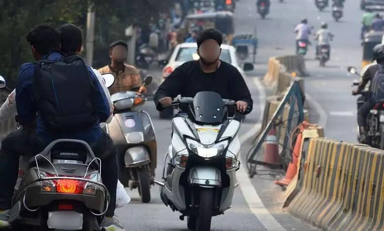 Scooter riders and pedestrians on busy urban street with traffic and road barriers in the background.