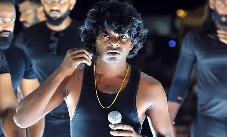 Young male singer with curly hair and black tank top singing into a microphone during a live performance.