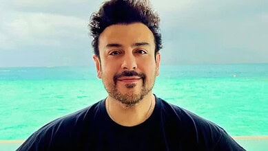 A man with dark hair and a beard smiling at the camera on a beach with turquoise water and a cloudy sky.