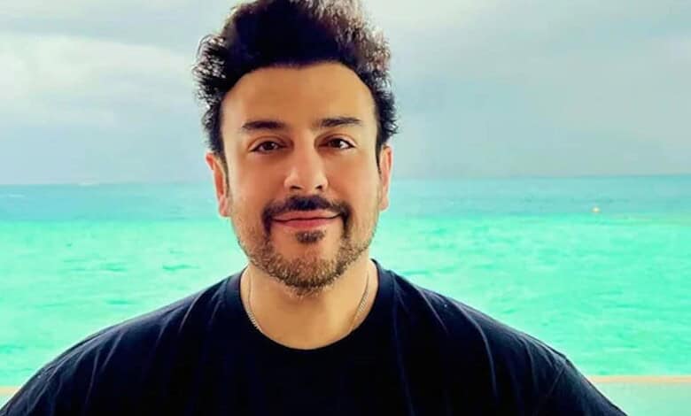 A man with dark hair and a beard smiling at the camera on a beach with turquoise water and a cloudy sky.
