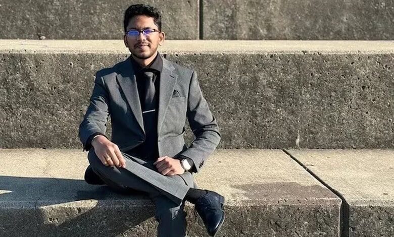 Young man in formal attire sitting on a concrete bench outdoors, wearing glasses and holding a tablet.