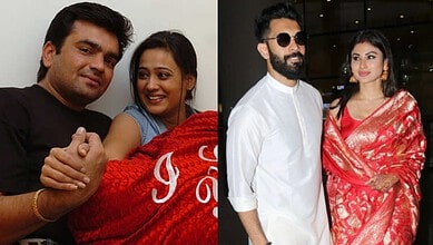 A man and woman holding hands, smiling, with the woman wearing a red saree.