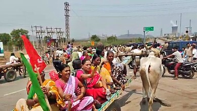 A group of women and children riding a bullock cart on a busy roadside, with vehicles and people in the background.