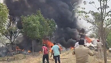Vehicle on fire with thick black smoke during an outdoor incident, surrounded by trees and onlookers in t.