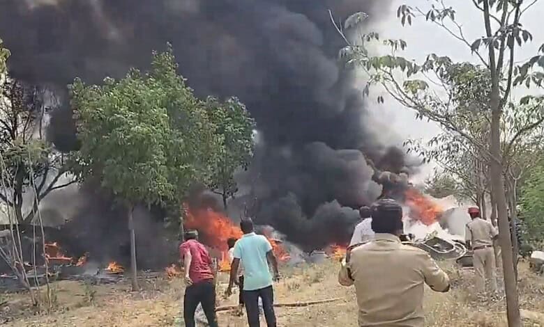 Vehicle on fire with thick black smoke during an outdoor incident, surrounded by trees and onlookers in t.