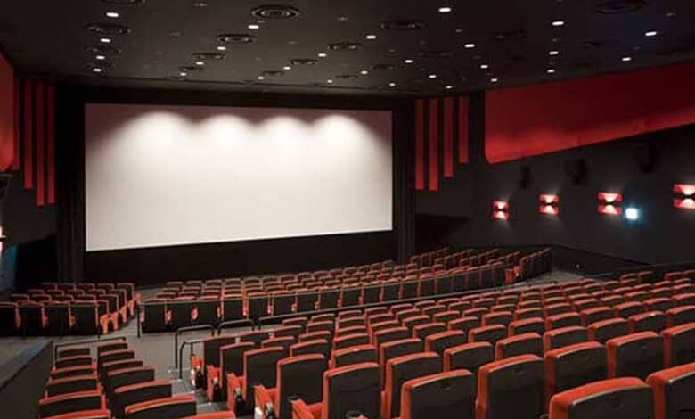 A nearly empty movie theatre with a large blank screen and rows of red and black seats.