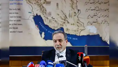 Indian politician speaking at a press conference with microphones, backdrop of a map of Iran and surround.