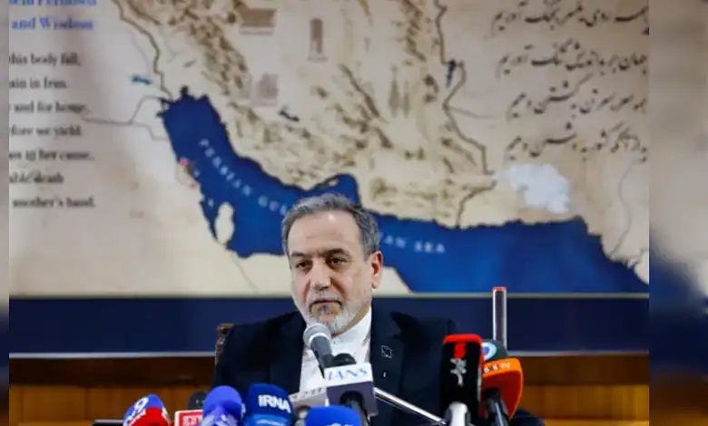 Indian politician speaking at a press conference with microphones, backdrop of a map of Iran and surround.