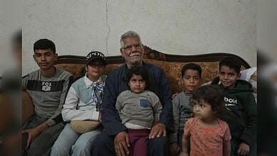 Elderly man with white hair and glasses sitting on a sofa surrounded by six children, in a simple indoor.