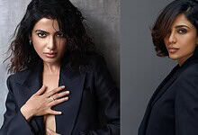 Elegant woman with wavy hair wearing a dark blazer, posing confidently against a grey background.