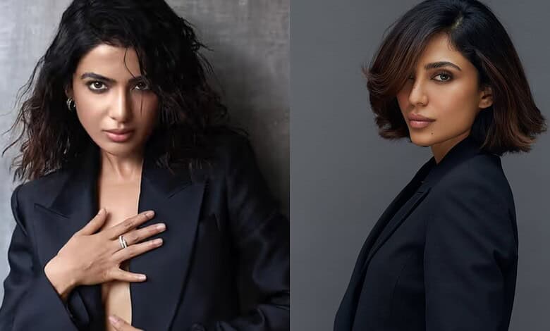 Elegant woman with wavy hair wearing a dark blazer, posing confidently against a grey background.