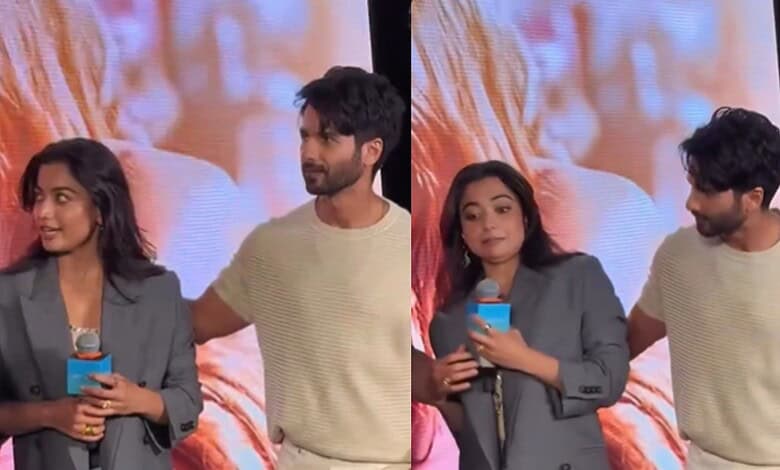 A woman and a man on stage during a media event, with a large screen displaying an image in the backgroun.