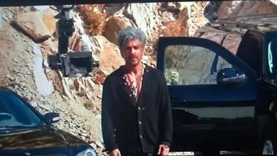 A man standing outdoors near a black car with a rocky background.
