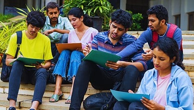 Group of diverse students sitting on steps, studying and discussing with notebooks and tablets.