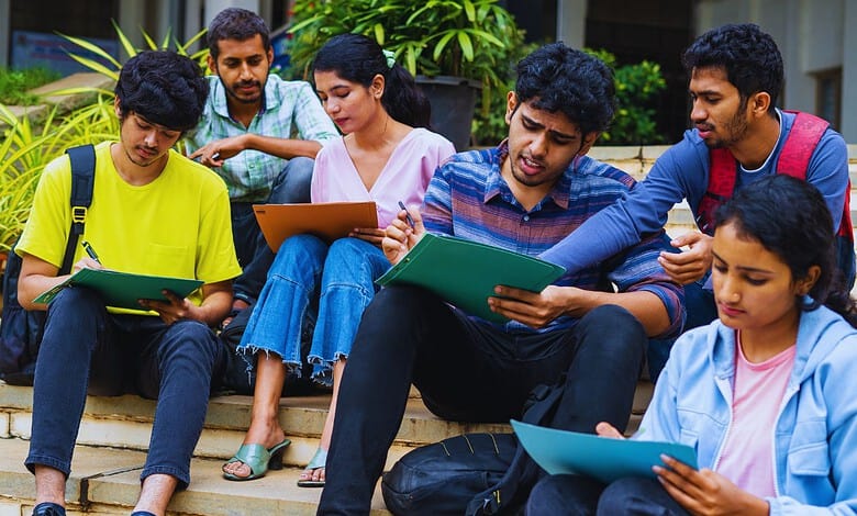 Group of diverse students sitting on steps, studying and discussing with notebooks and tablets.