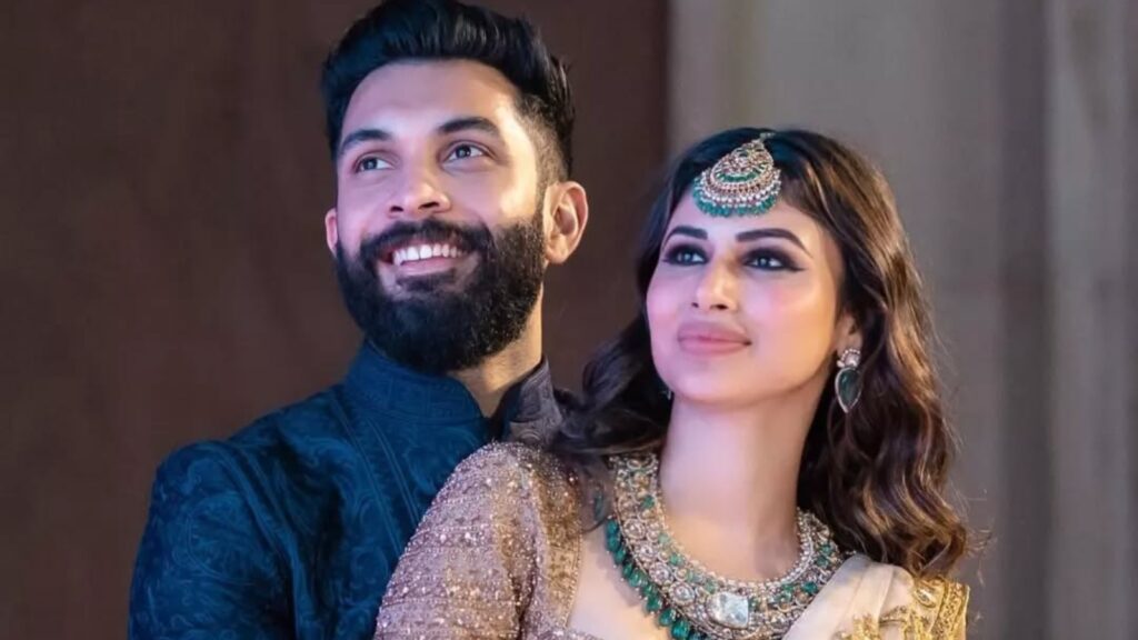 A woman and a man smiling, dressed in traditional Indian attire, with a neutral background.