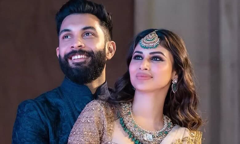 A woman and a man smiling, dressed in traditional Indian attire, with a neutral background.