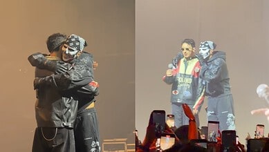 A woman and a man hugging on stage during a music event, with another performer speaking into a microphon.