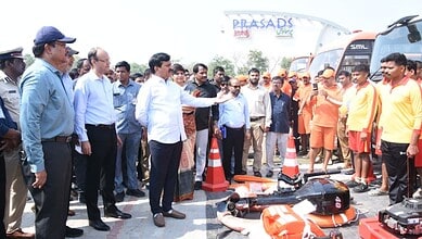 A man demonstrating safety equipment to a group of people at an outdoor event with traffic cones and a bu.