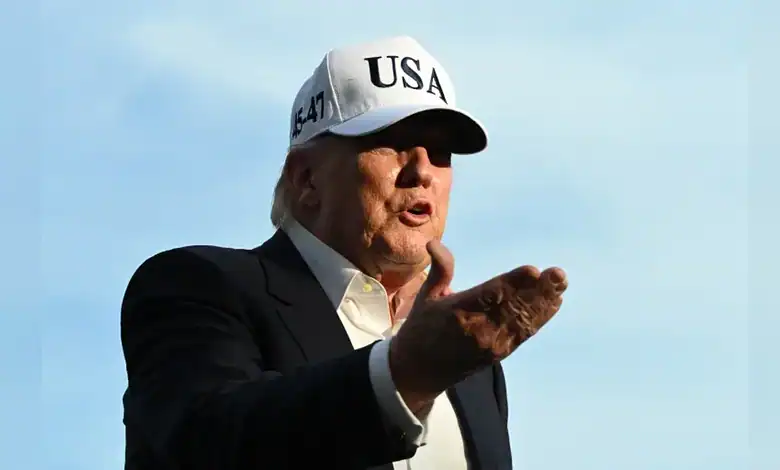 Trump in a "USA" cap, speaking outdoors with a blue sky, making a gesture during a public appearance.